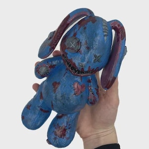 May include: A blue plush bunny with long ears and a stitched mouth. The bunny has red accents and button eyes. The bunny is held in a hand against a white background.