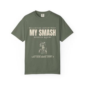 May include: An olive green short-sleeve t-shirt featuring cream-coloured text that reads BALLISTICS DEPARTMENT MY SMASH REPORTED MISSING LAST SEEN ABOVE COURT 3, accompanied by a small, detailed line-art illustration of a tennis player in motion.