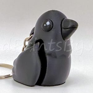 May include: A dark gray crow-shaped keychain. The crow has a rounded body, a small beak, and large, round eyes. The keychain ring is silver. The text "Cute 3d Prints by John" is visible in the background, along with a seller's logo.