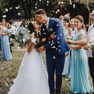 Op de afbeelding: Een bruidspaar kust elkaar terwijl er witte bloemblaadjes naar beneden vallen. De persoon links draagt een witte kanten jurk en houdt een blauw-wit boeket vast. De persoon rechts draagt een marineblauw pak met een wit overhemd en das.