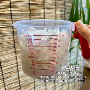Op de afbeelding: Een doorzichtige plastic maatbeker met een rood handvat gevuld met witte bloemblaadjes. De beker heeft rode maataanduidingen in cups en ounces, met de tekst 1 QUART SUFFICIENT FOR: 20 SINGLE HANDFUL TOSSES 10 DOUBLE HANDFUL TOSSES.