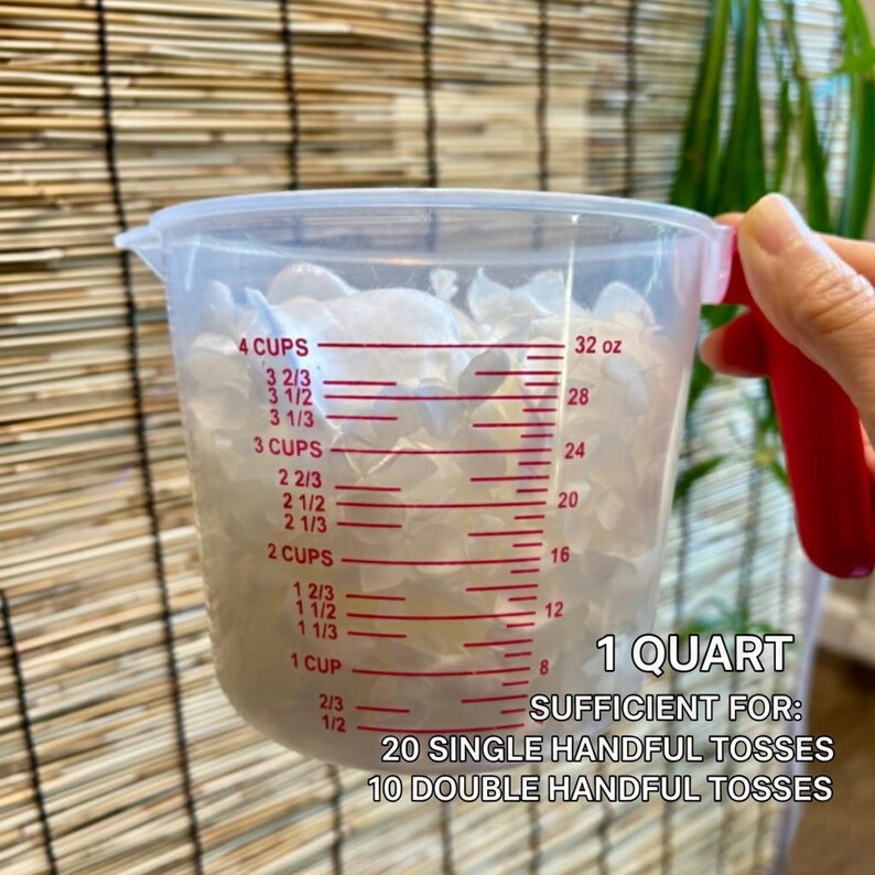 Op de afbeelding: Een doorzichtige plastic maatbeker met een rood handvat gevuld met witte bloemblaadjes. De beker heeft rode maataanduidingen in cups en ounces, met de tekst 1 QUART SUFFICIENT FOR: 20 SINGLE HANDFUL TOSSES 10 DOUBLE HANDFUL TOSSES.