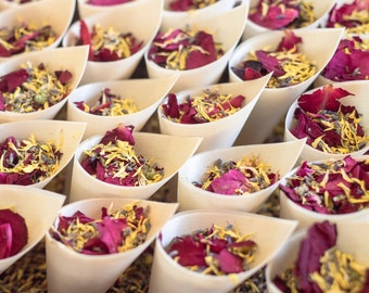 May include: A collection of small white paper cones filled with dried flower petals in shades of pink, yellow, and lavender. The cones are arranged in rows on a woven surface.