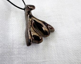 Könnte beinhalten: Eine Halskette mit einem schwarzen Keramik-Anhänger in einem einzigartigen, abstrakten Design. Der Anhänger ist in Form einer Gruppe von drei abgerundeten Formen gestaltet, die an eine stilisierte Blume oder eine Traube erinnern. Die Halskette ist aus einem schwarzen Band gefertigt.
