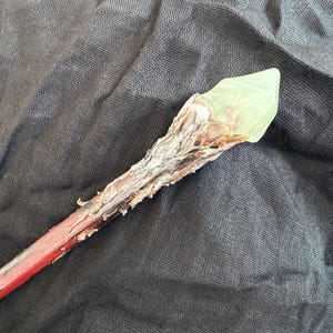 Può includere: Una bacchetta fatta a mano con un'impugnatura rossa e un fusto strutturato di colore chiaro. La punta della bacchetta presenta un cristallo verde chiaro traslucido. La bacchetta è posta su uno sfondo scuro e strutturato.