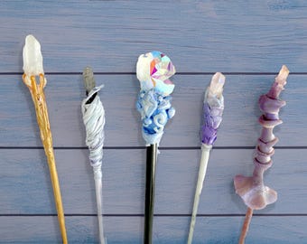 Puede incluir: Cinco varitas hechas a mano con diseños únicos. Una varita tiene una parte superior de cristal transparente y detalles dorados. Otra está envuelta en espirales blancas y grises. Otras varitas presentan partes superiores de cristal azul, morado y rosa.
