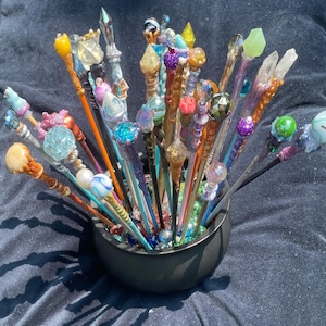 Può includere: Una collezione di bacchette decorative con cime colorate a forma di gemma e disegni intricati. Le bacchette sono esposte in una ciotola nera, mostrando una varietà di colori e trame, tra cui cristalli trasparenti e perle iridescenti.