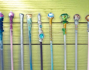 Puede incluir: Una colección de nueve varitas hechas a mano con diseños únicos. Cada varita presenta una combinación de colores diferente y una parte superior decorativa, que incluye remolinos, gemas y elementos esculpidos. Las varitas están dispuestas sobre una superficie de madera verde claro.