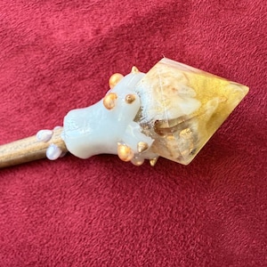 May include: A handcrafted wand with a light wood handle and a decorative head. The head features a white, sculpted element adorned with gold accents and a clear, gold-infused resin tip. The wand rests on a red surface.