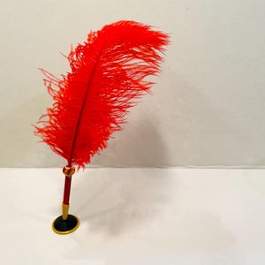 Op de afbeelding: Een decoratieve ganzenveerpen met een felrode veer. De pen heeft een donkerrode schacht met gouden en zwarte details. De veer is lang en pluizig, wat bijdraagt aan het sierlijke ontwerp van de pen.
