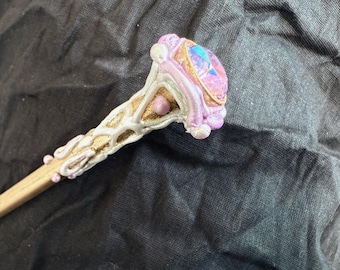 May include: A handcrafted wooden wand with a decorative head. The head features a large, iridescent gemstone set in a gold-colored frame, surrounded by white and pink embellishments. The wand's shaft is light brown.