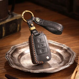 May include: Black leather car key fob cover with orange stitching and a metal clasp. The fob has three buttons and a small silver button. The key fob is attached to a black leather strap with the text "GB-0303".