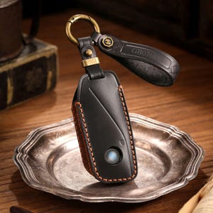 May include: Black leather key fob holder with a brown stitched edge and a metal clasp. The fob has a circular blue light and a black leather strap with the text "GB07688". The key fob is on a wooden surface.