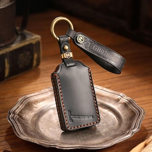 May include: Black leather key fob with a gold-colored ring and accents. The fob has a stitched border and a strap with the text "GB078669". The key fob is resting on a silver-colored plate.