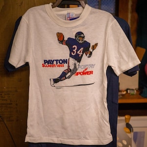 May include: White t-shirt featuring a football player in a navy blue uniform with the number 34. The shirt has the words "Payton Sweetness Poetry Power" printed in red and blue.