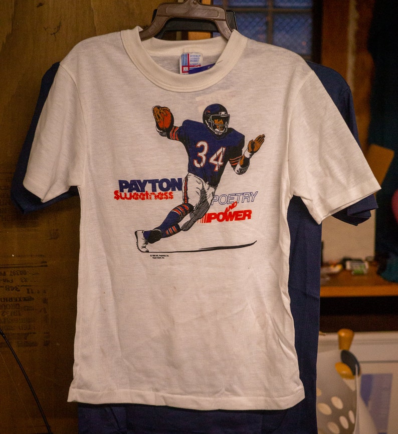 May include: White t-shirt featuring a football player in a navy blue uniform with the number 34. The shirt has the words "Payton Sweetness Poetry Power" printed in red and blue.