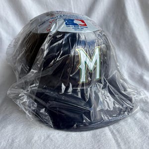 Op de afbeelding: Een marineblauwe baseballcap met een wit en groen "M" logo. De cap zit in een doorzichtige plastic zak. De verpakking heeft tekst en een Major League Baseball logo.