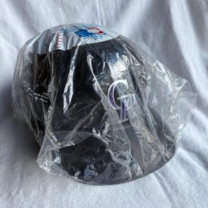 Op de afbeelding: Een zwarte baseballcap met het logo van de Colorado Rockies in wit en paars. De cap zit in de originele verpakking, een doorzichtige plastic zak. De bovenkant van de cap heeft een wit gedeelte met rode stiksels.