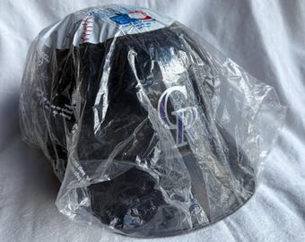 May include: A black baseball cap with the Colorado Rockies team logo in white and purple. The cap is still in its original packaging, a clear plastic bag. The top of the cap has a white section with red stitching.