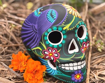 May include: A black ceramic skull sculpture featuring intricate hand-painted designs in vibrant colors including purple, blue, green, yellow, and pink. The skull displays floral patterns, a bird motif, and white teeth, with two bright orange marigold flowers.