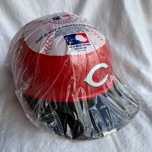 Op de afbeelding: Een Cincinnati Reds honkbalhelm in een doorzichtige plastic zak. De helm is rood met een wit "C"-logo en een zwarte klep. De bovenkant van de helm heeft een honkbalontwerp en de tekst "THIS IS NOT A PROTECTIVE HELMET".