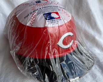 May include: A Cincinnati Reds baseball helmet in a clear plastic bag. The helmet is red with a white "C" logo and a black brim. The top of the helmet has a baseball design and text that reads "THIS IS NOT A PROTECTIVE HELMET".