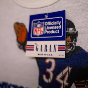 May include: White t-shirt featuring a football player in a blue and white uniform with the number 34. The shirt has a tag that reads "Officially Licensed Product" by Garan, Made in U.S.A. NFL-20.