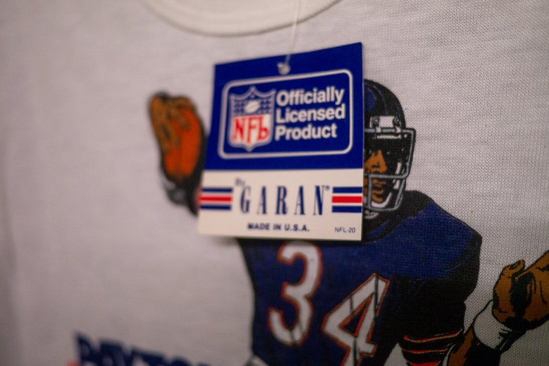 May include: White t-shirt featuring a football player in a blue and white uniform with the number 34. The shirt has a tag that reads "Officially Licensed Product" by Garan, Made in U.S.A. NFL-20.