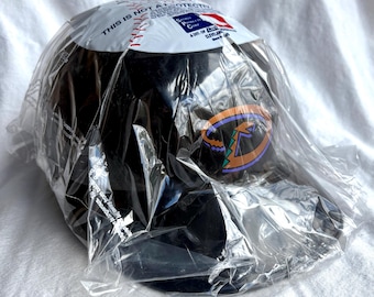 May include: A black baseball cap with a team logo in purple, orange, and green. The cap is packaged in clear plastic. The top of the cap has the text "THIS IS NOT A PROTECTIVE".