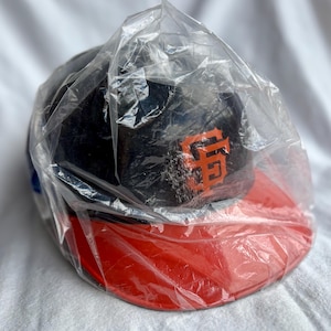 Op de afbeelding: Een marineblauwe baseballcap met een oranje SF-logo op de voorkant en een rode klep. De cap is verpakt in een doorzichtige plastic zak. De cap is waarschijnlijk een merchandise-artikel van een sportteam.