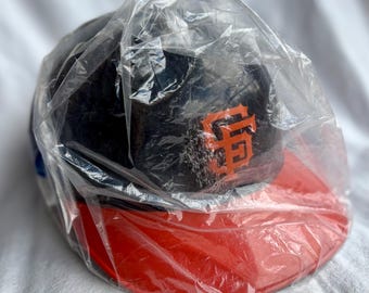 May include: A navy blue baseball cap with an orange SF logo on the front, and a red brim. The cap is encased in a clear plastic bag. The cap is likely a sports team merchandise item.