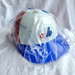 Op de afbeelding: Een baseballpet met een witte kroon, rood-witte zijpanelen en een blauwe klep. De pet heeft een gestileerd ontwerp met een rode cirkel en een blauwe vorm. Verpakt in een doorzichtige plastic zak.