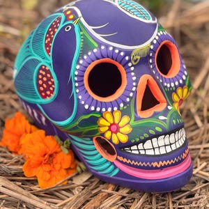 May include: A purple skull sculpture featuring hand-painted details including bright orange eye sockets and nasal cavity, yellow flowers, white teeth, and teal bird motifs. The surface is decorated with intricate patterns in pink, green, and white.