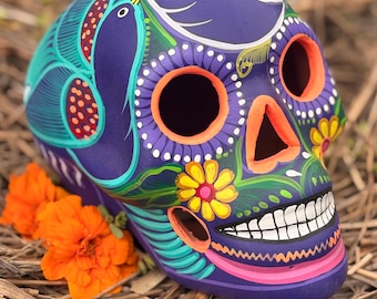 May include: A purple skull sculpture featuring hand-painted details including bright orange eye sockets and nasal cavity, yellow flowers, white teeth, and teal bird motifs. The surface is decorated with intricate patterns in pink, green, and white.