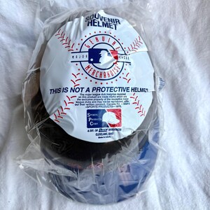 Op de afbeelding: Een souvenir honkbalhelm in een doorzichtige plastic zak. De helm is zwart en blauw met een wit label met de tekst "Souvenir Helmet" en "Genuine Merchandise". Op het label staat ook "This is not a protective helmet."
