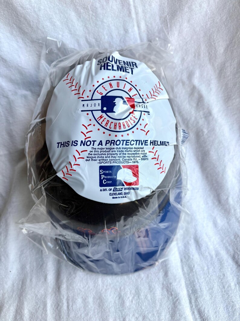 Op de afbeelding: Een souvenir honkbalhelm in een doorzichtige plastic zak. De helm is zwart en blauw met een wit label met de tekst "Souvenir Helmet" en "Genuine Merchandise". Op het label staat ook "This is not a protective helmet."