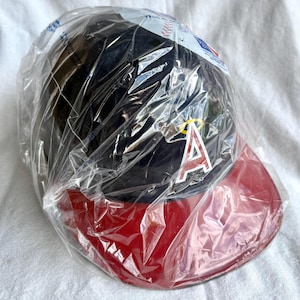Op de afbeelding: Een zwarte baseballcap met een rode klep, verpakt in doorzichtig plastic. De cap heeft een rood en wit "A" logo met een gele halo. Dit is waarschijnlijk een merchandise-artikel van een sportteam.