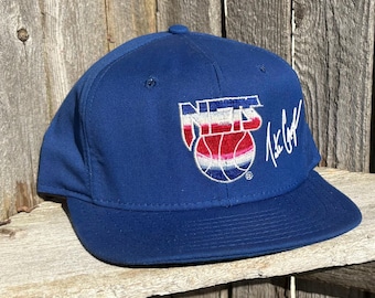 May include: A royal blue baseball cap featuring an embroidered Nets logo in red, white, and blue with a basketball graphic. The cap includes a white embroidered signature on the right side and has a structured crown with a flat brim and eyelets.