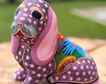May include: A hand-painted dog figurine featuring a purple base with pink polka dots, long solid pink ears, and a black back decorated with vibrant floral patterns in red, yellow, blue, and green. The dog has black eyes and white whiskers.
