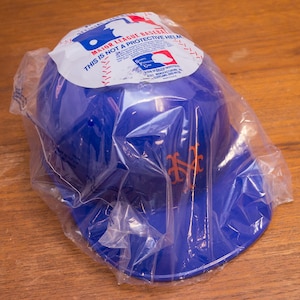 Puede incluir: Una gorra de b&eacute;isbol de pl&aacute;stico azul con el logotipo de los Mets de Nueva York en naranja. La gorra est&aacute; empaquetada en una bolsa de pl&aacute;stico transparente con una etiqueta de advertencia que dice "Major League Baseball. Esto no es un casco protector."
