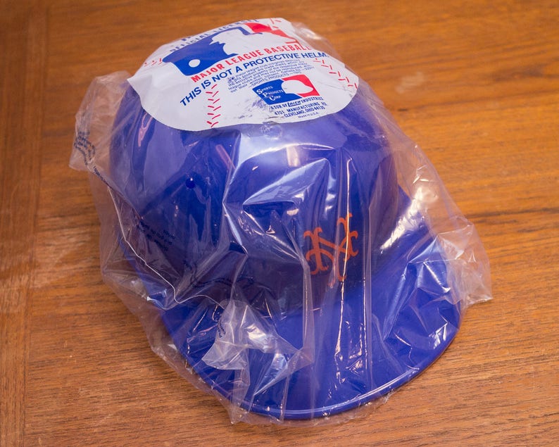 Puede incluir: Una gorra de b&eacute;isbol de pl&aacute;stico azul con el logotipo de los Mets de Nueva York en naranja. La gorra est&aacute; empaquetada en una bolsa de pl&aacute;stico transparente con una etiqueta de advertencia que dice "Major League Baseball. Esto no es un casco protector."