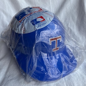 Op de afbeelding: Blauwe honkbalhelm met een rood "T" logo, verpakt in doorzichtig plastic. De helm heeft de tekst "MAJOR LEAGUE BASEBALL" en "THIS IS NOT A PROTECTIVE HELMET".