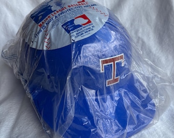 May include: A blue baseball helmet with a red "T" logo, packaged in clear plastic. The helmet has text that reads "MAJOR LEAGUE BASEBALL" and "THIS IS NOT A PROTECTIVE HELMET".