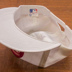 May include: White baseball cap with a red logo that says "Cardinals" on the side. The cap has a white adjustable strap at the back.