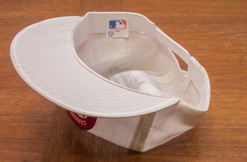 May include: White baseball cap with a red logo that says "Cardinals" on the side. The cap has a white adjustable strap at the back.