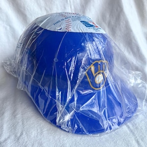 Op de afbeelding: Een blauwe baseballpet met het Milwaukee Brewers-logo in geel en wit, in een doorzichtige plastic verpakking. De bovenkant van de pet heeft een witte band met rode honkbalstiksels en tekst.