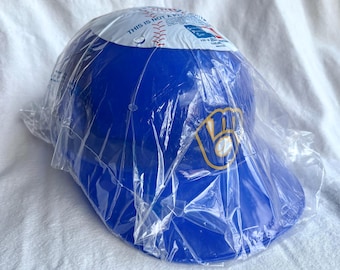 May include: A blue baseball cap with the Milwaukee Brewers logo in yellow and white. The cap is in a clear plastic wrapper. The top of the cap has a white band with red baseball stitching and text.