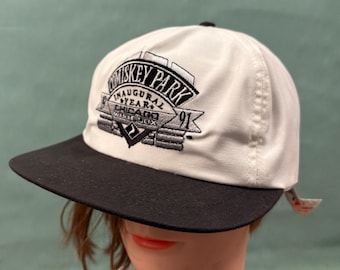 May include: A white baseball cap with a black brim featuring black and grey embroidery on the front. The text reads Comiskey Park Inaugural Year 1991 Chicago White Sox with a graphic of a stadium structure and a diamond shape logo below the text.