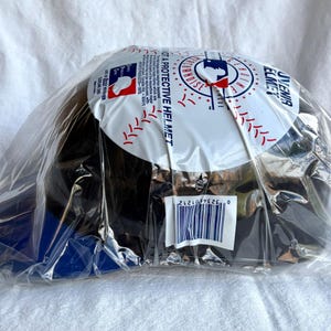 Op de afbeelding: Een zwarte baseballpet met een blauwe klep, verpakt in een doorzichtige plastic zak. De bovenkant van de pet heeft een wit cirkelvormig ontwerp met rode en blauwe accenten en de tekst "A PROTECTIVE HELMET".