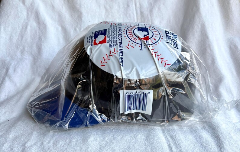 Op de afbeelding: Een zwarte baseballpet met een blauwe klep, verpakt in een doorzichtige plastic zak. De bovenkant van de pet heeft een wit cirkelvormig ontwerp met rode en blauwe accenten en de tekst "A PROTECTIVE HELMET".